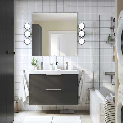 Moderne badkamer met zwart HAVBÄCK badmeubel, witte dubbele wastafel, grijze tegels en ronde spiegels. Witte handdoeken, zwarte wasdroger.