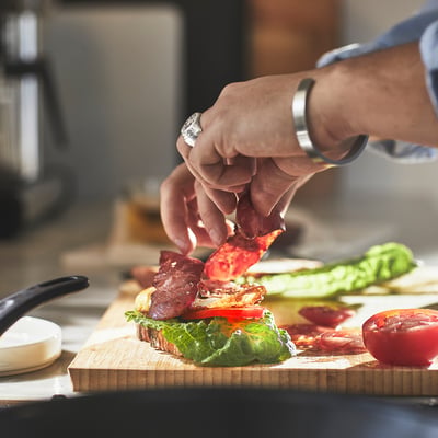 Handen die spek toevoegen aan sandwich op houten plank naast tomaten.