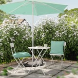 Kleine buitentafel met lichtgroene stoelen met kussens en mint parasol, omgeven door bloeiende struiken.