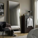 Grote HOVET-spiegel met gouden lijst weerspiegelt de slaapkamer en verbetert de ruimte. Gepositioneerd naast een hoog dressoir, domineert het de kamer, het reflecterende raam en het decor.