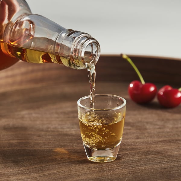 Een transparant glas is gevuld met vloeistof uit een fles, waarschijnlijk gebruikt voor het proeven van sterke dranken zoals baijiu of sake. Het glas staat op een houten tafel.