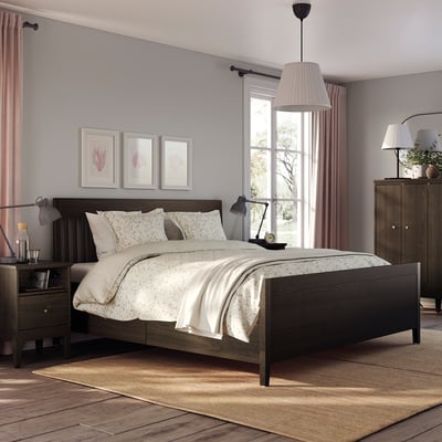 Slaapkamer met IDANÄS donkerbruin bed, wit beddengoed, nachtkastjes en gordijnen; groot raam, vloerkleed en potplant.