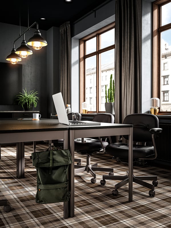 Moderne kantoorruimte met donkere meubels: IDÅSEN tafels, zwarte stoelen, groene rugzak, planten, hanglampen.