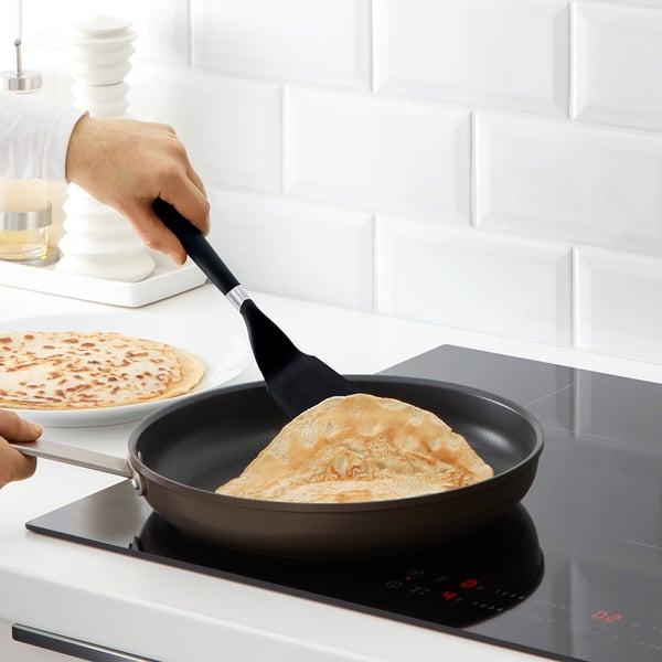 Persoon gebruikt ikea-spatel om pannenkoek in een koekenpan op een inductiefornuis om te draaien. Witte keuken achtergrond.