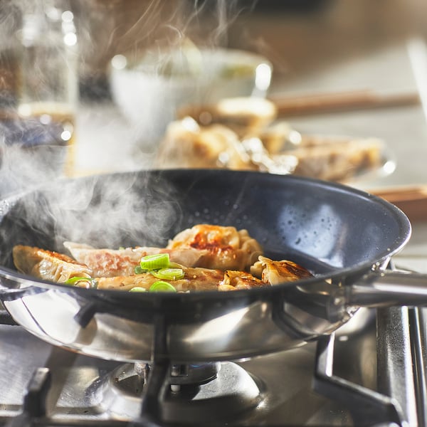Afbeeldingsbeschrijving: een zwarte koekenpan op een fornuis, kookknoedels en groenten. Stoom stijgt op, wat wijst op actief koken.
