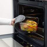 Persoon die glazen voedselcontainer, gevuld met groenten, in een open oven plaatst met behulp van een ovenwant.