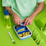 De persoon in groen overhemd opent blauwe lunchbox die voedsel op groene doek bevat. Een blauwe waterfles met een handvat is in de buurt, samen met een witte vork.