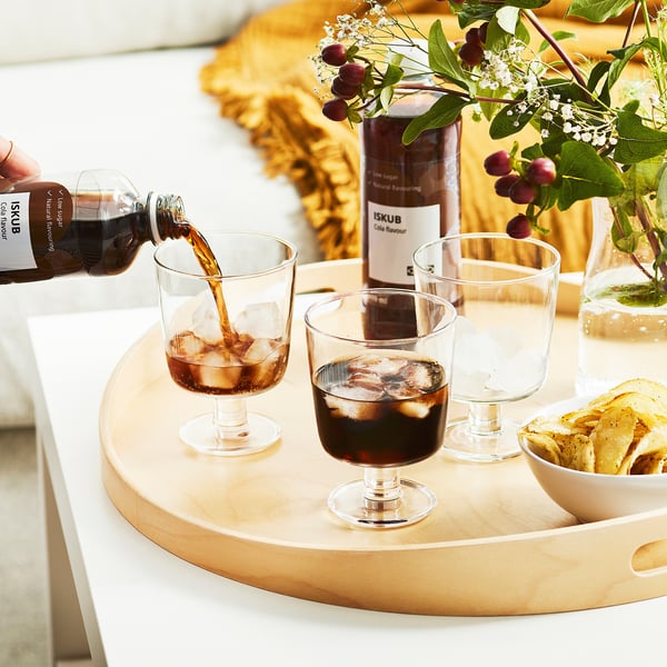 Drie glazen op een houten dienblad ontvangen cola uit een fles, met op de achtergrond een schaal met snacks en een vaas met bloemen.