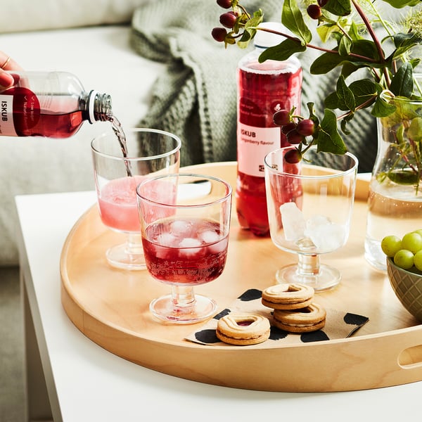 Persoon die roze drank in een glas op een houten dienblad giet dat koekjes, druiven en een fles met het label ISKUB-lingonberry-smaak bevat.