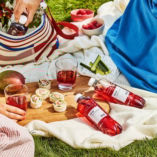 Picknickscène met ISKUB-drankjes, broodjes, fruit en snacks op een deken. Iemand schenkt ISKUB-drank in een glas.