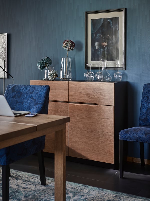 Moderne eethoek met KLACKENÄS eiken dressoir, stoelen met blauwe patronen, glaswerk en abstracte kunst aan de muur op blauwe muur.