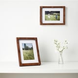 Twee fotolijsten tonen met gras begroeide scènes; De een staat, de ander hangt met een vaas.