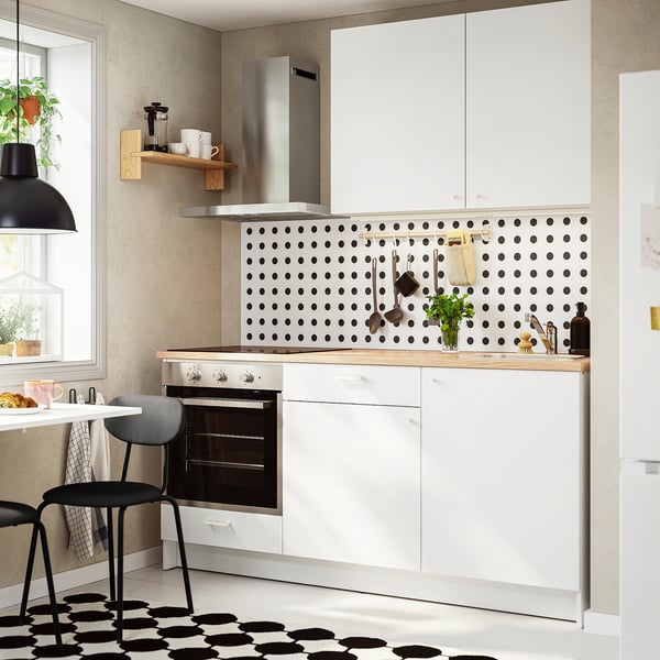 Moderne witte keukenopstelling. Zwarte achterwand met stippen, houten plank, kleine oven, aanrechtaccessoires. Compact ontwerp past goed in kleinere keukens.