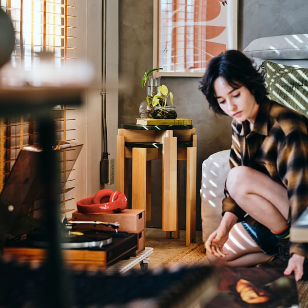 Persoon met baby op een patroonbank, houten KOMPISHÄNG-krukjes als plantenstandaard, rood speelgoed op de vloer.