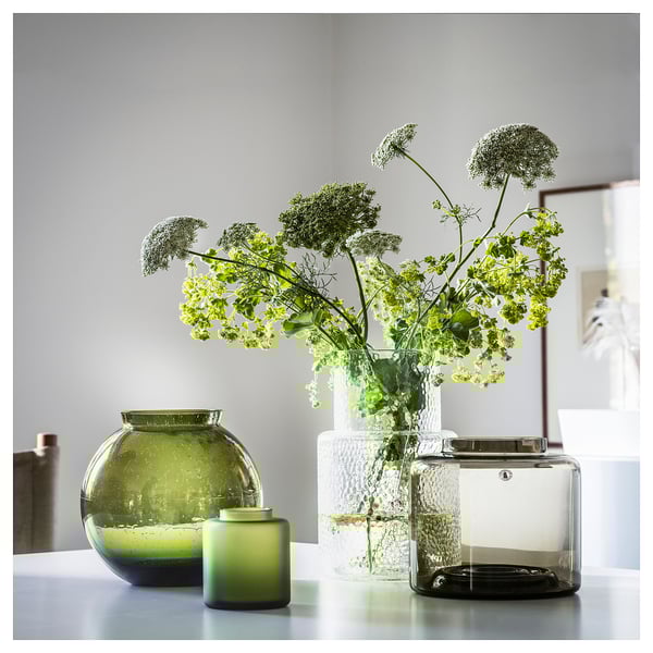 Drie vazen op een tafel: een groene, een heldere, een mat. Groene vaas bevat witte bloemen. Heldere vaas is lang, mat is rond.