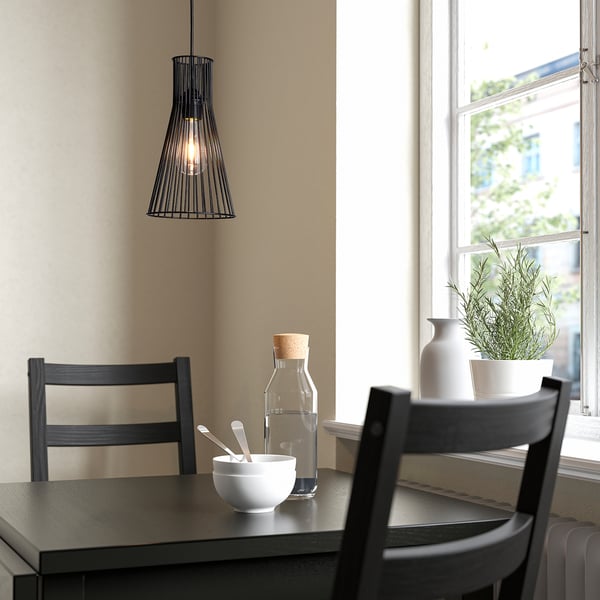 Boven een donkerhouten tafel hangt een kleine hanglamp van zwart draad naast twee donkerhouten stoelen bij een raam met een wit frame.