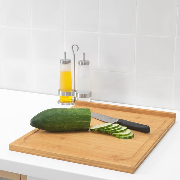 Houten snijplank met mes en gesneden komkommer; oliedispensers achter.