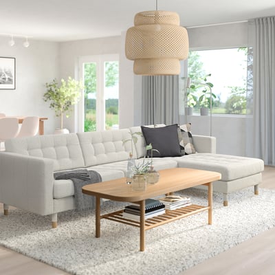 Moderne woonkamer met LANDSKRONA sectionaal, houten tafel, rieten lamp, vloerkleed, planten en ramen.
