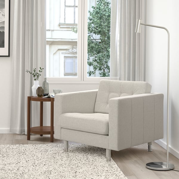 Een beige LANDSKRONA fauteuil bij een groot raam, met daarnaast een vloerlamp en een decoratieve tafel.