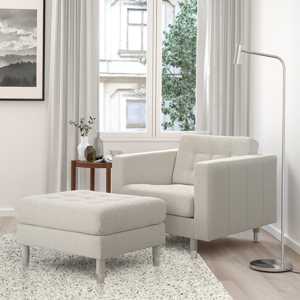 Moderne beige LANDSKRONA voetenbank naast fauteuil, met metalen poten en getuft design. Nabij raam met doorschijnende gordijnen en een bijzettafelvaas.