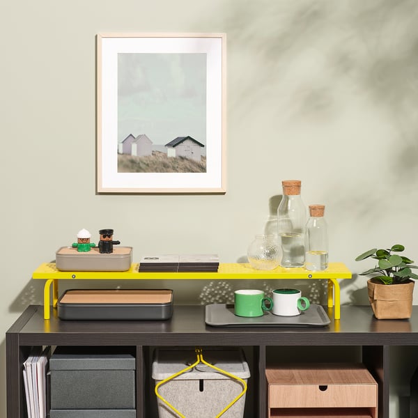 Een gele monitorstandaard tilt twee computers op voor een betere houding. Hieronder staan een lade, mokken en een plant op een plank.