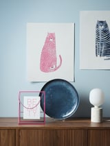 Een stijlvolle display op een donkerhouten plank: kleurrijke kattenprints, een blauw bord en een witte terrazzo-tafellamp.
