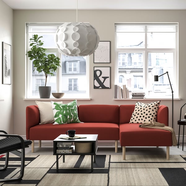 Moderne woonkamer met rode LILLEHEM bank, grote ramen, plant en zwarte salontafel.