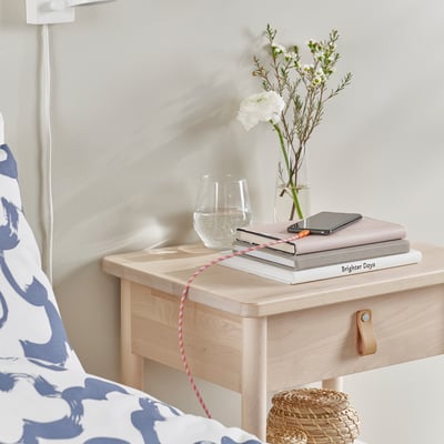 Nachtkastje naast het bed met een telefoon oplaadkabel, boeken, glas en bloemen.