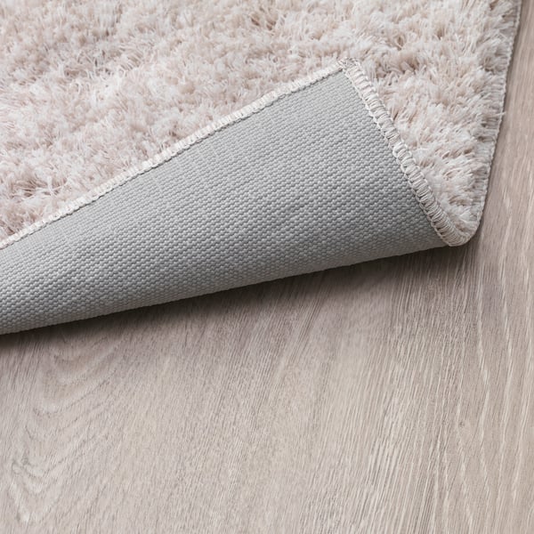 Een pluche vloerkleed gelegd op een houten vloer, met zachte beige vezels en een grijze antislip achterkant.