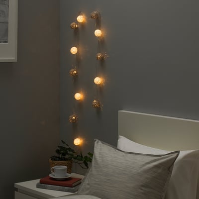 Gouden LIVSÅR-lampen op grijze muur. Warm bed met rood boek, beker en plant.