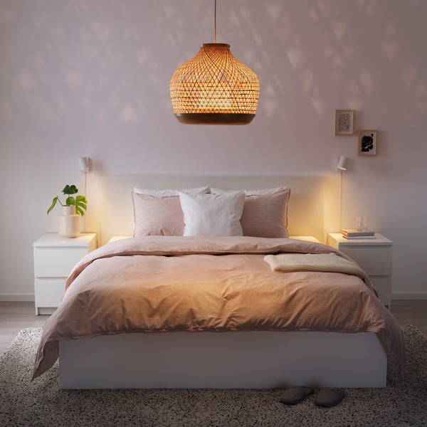 Gezellige slaapkamer met beige beddengoed, hangende rieten lamp, witte muren en kleine foto's. LUNNOM-lamp straalt warm licht uit. Slippers op het tapijt.