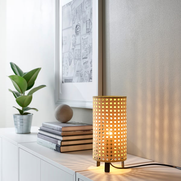 Een kleine bamboe tafellamp verlicht op de plank met boeken, plant en afbeelding.