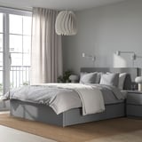 Modern grijs MALM bed met hoog hoofdeinde, grote lades voor opslag. Witte kussens en grijs linnengoed. Unieke hanglamp, groot raam met uitzicht.