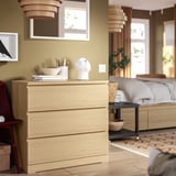 Houten MALM dressoir met decor en boeken. Bed met beige beddengoed, vloerkleed en groot raam.