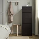 Moderne slaapkamer met MALM donkerbruin dressoir, wit bed, kruk, hanger met kleding en plant.