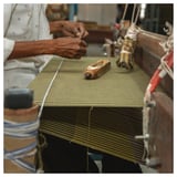 Persoon weeft geel gestreepte MÄVINN sprei op houten weefgetouw. Levendige strepen, katoenen materiaal, handwerk duidelijk.