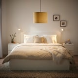 Slaapkamer met grote, ronde gele MOLNSKIKT lampenkap, zachte verlichting, wit bed, twee nachtkastjes en ingelijste foto's aan de muur.