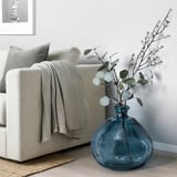 Beige bank met grijze deken, blauwe gerecycleerde glazen vaas MYRMOSAIK op houten vloer met decoratieve takken.