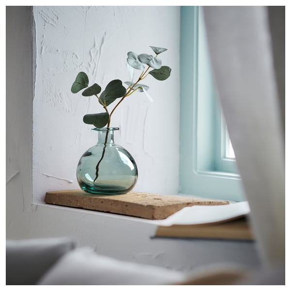 Kleine glazen vaas met groen-blauwe tint op rustieke houten plank. Bevat enkelstammige eucalyptus.