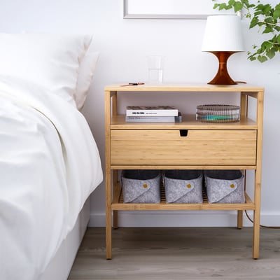 Klein licht bamboe nachtkastje met een lade en open planken. Het past netjes naast een bed, met een lamp, boeken en opbergbakken.