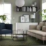 Moderne woonkamer met saliegroene muren, bruine bank, blauwe stoel, groen vloerkleed, glazen tafel en planten.