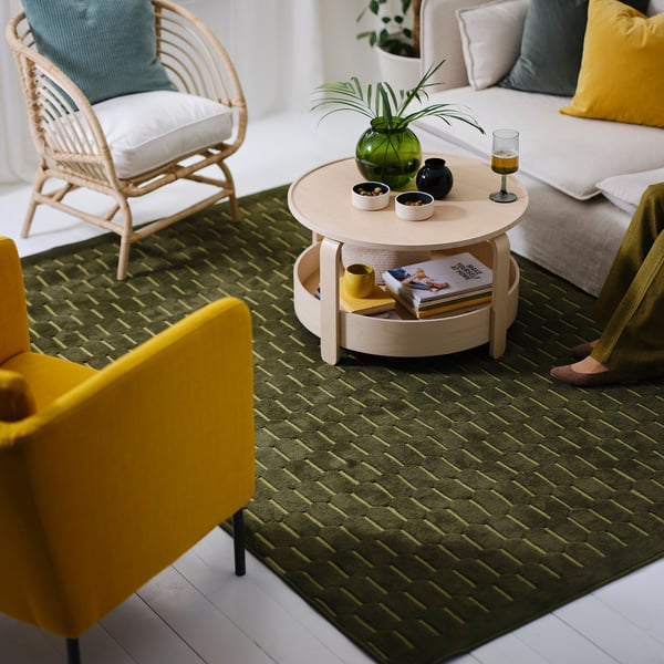 Woonkamer met groen NÖVLING rugby, lichthouten salontafel, felgele stoel, witte bank met kleurrijke kussens en een groene vaas.
