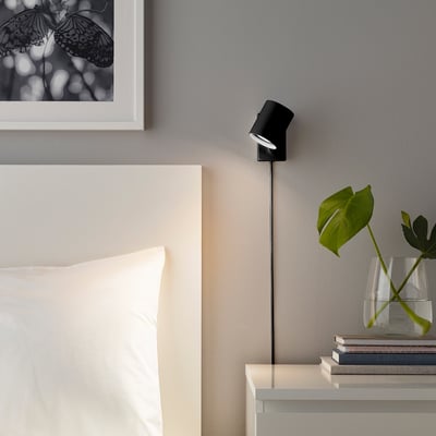 Aan de muur gemonteerde verstelbare lamp, NYMÅNE, naast het bed, straalt warm licht uit, in de buurt van de plant en de fotolijst.