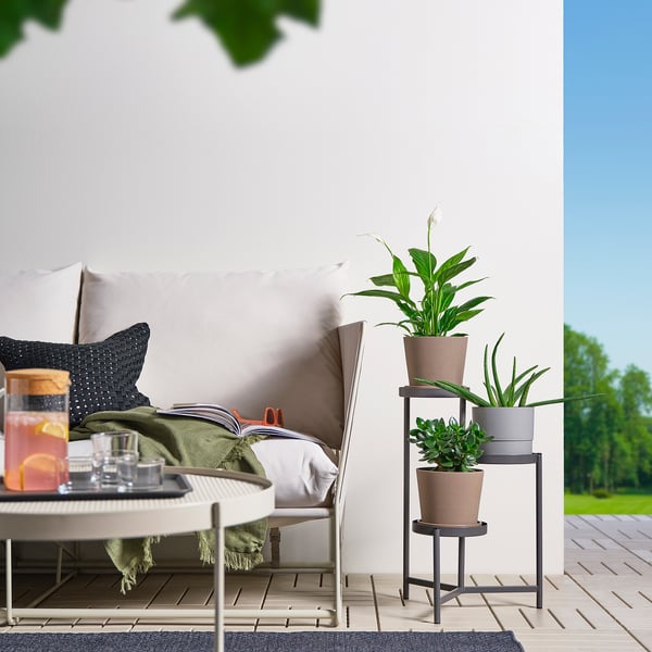 Moderne patio met grijze OLIVBLAD plantenstandaard, potplanten, kussenstoel, tafel met drankje. Gezellige sfeer, witte muur, groen.