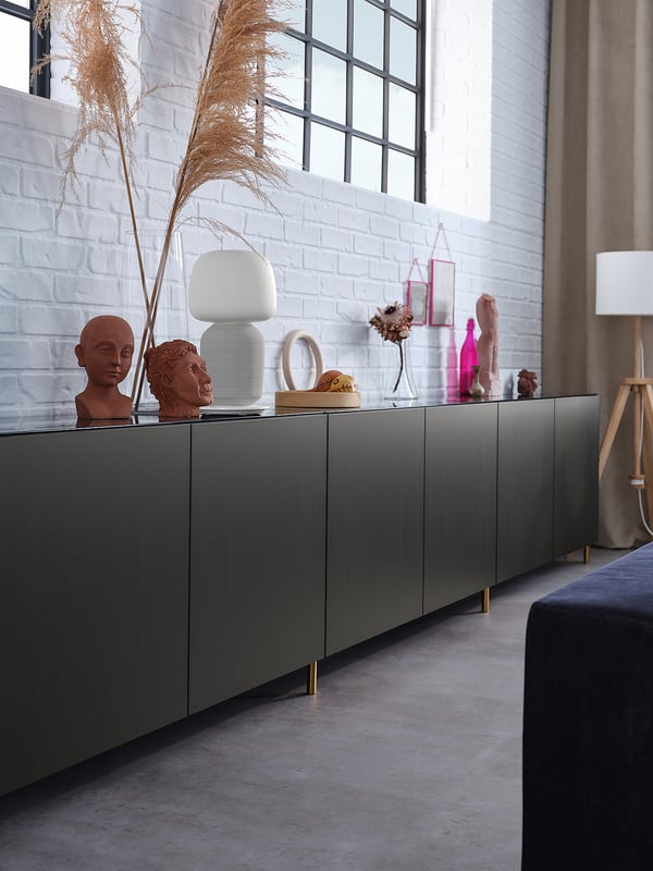 Een moderne woonkamer met een groen dressoir versierd met sculpturen, kaarsen en een lamp. Witte bakstenen muur, grote ramen en een lamp in de hoek.