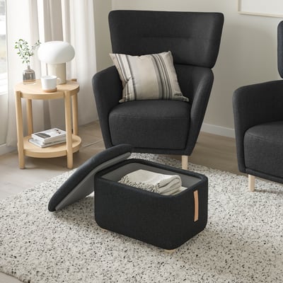 Moderne woonkamer met grijze stoel met hoge rugleuning, gestreept kussen, houten bijzettafel, lamp, mok en open voetenbankje waarin een deken kan worden opgeborgen.