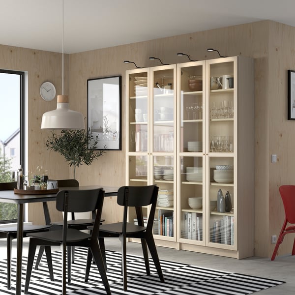 Moderne eethoek met OXBERG boekenkast, houten tafel, zwarte stoelen en gestreept vloerkleed. Potplant en wandklok versterken het decor.