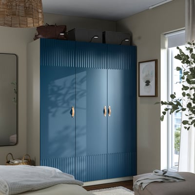 Moderne slaapkamer met een hoge blauwe kledingkast met soft-close deuren en opbergdozen. Geflankeerd door een spiegel en plant.