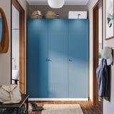 Grote blauwe PAX kledingkast domineert de kamer, hoge houten deuren, bovenste manden, kapstokken aan de rechterkant, lamp op tafel aan de linkerkant.
