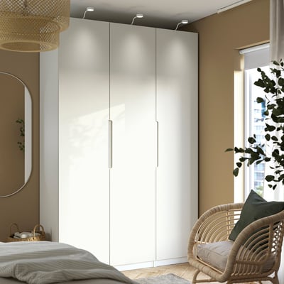 Moderne slaapkamer met hoge PAX kledingkast, ronde spiegel, rieten stoel en groot raam.